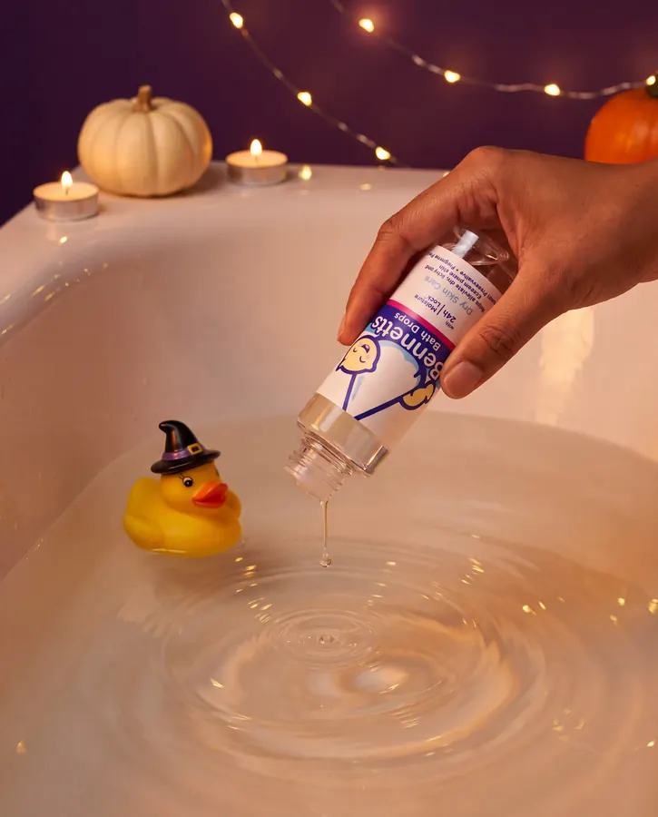 A hand pouring Bennetts Bath Drops into a bath beside a witch-hat rubber duck and tealights